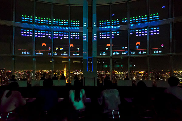 Visitors playing Space Invaders Gigamax projected across large windows above the Tokyo night skyline